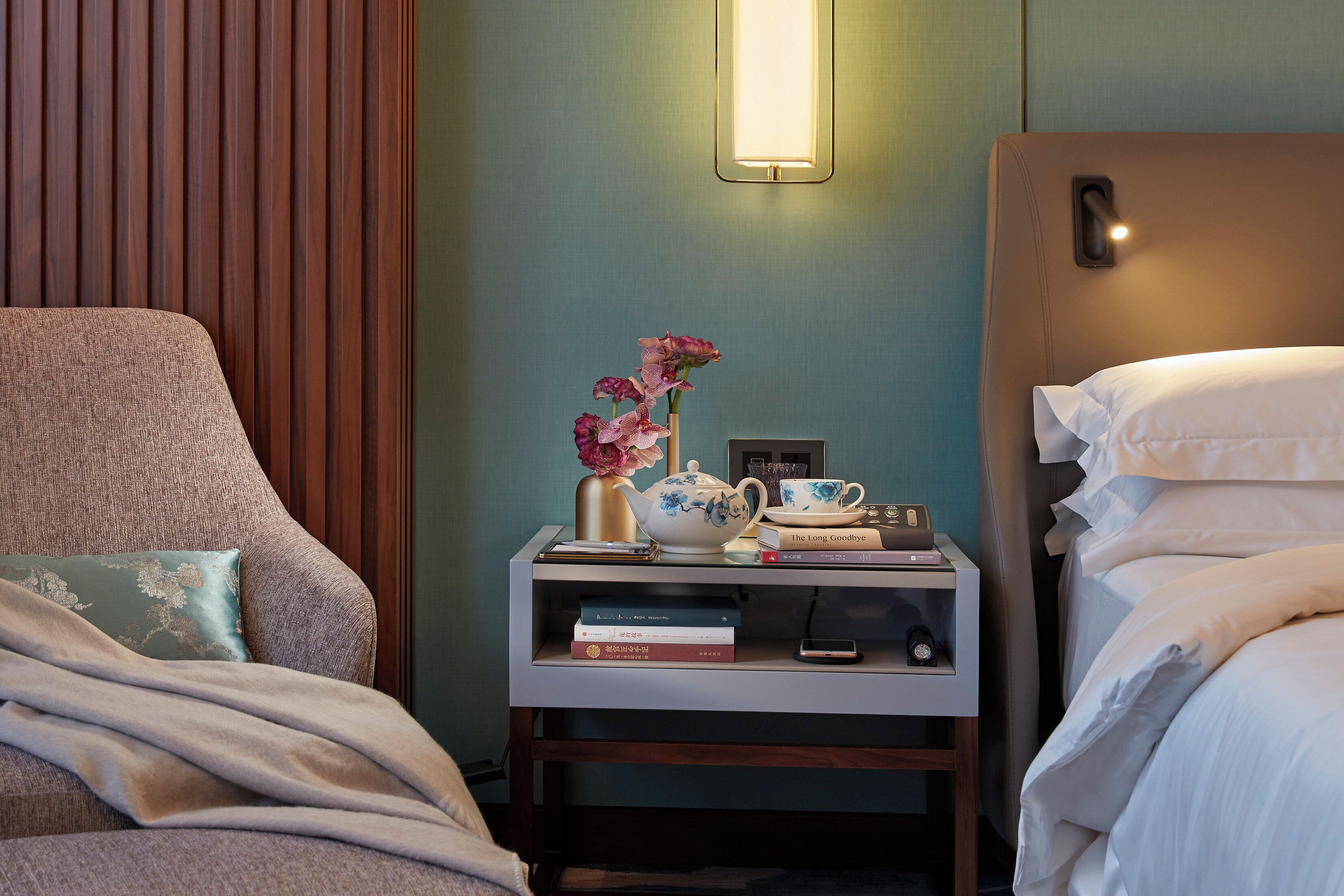 A neatly arranged hotel room with a bed, a chair, a bedside table with books and a teapot, and a wall-mounted light fixture.