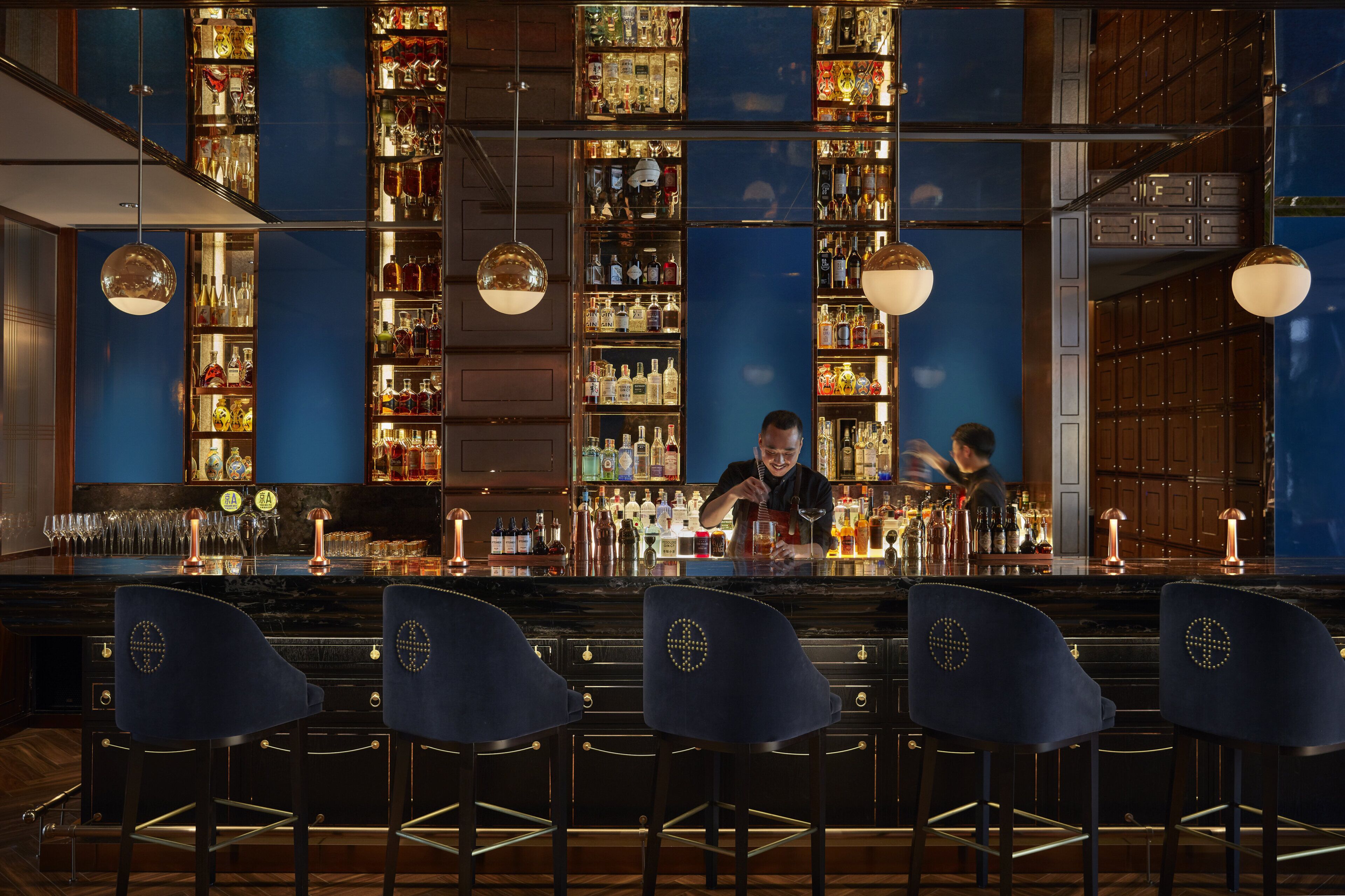 A bar with multiple shelves of bottles, a bartender preparing drinks, and bar stools.