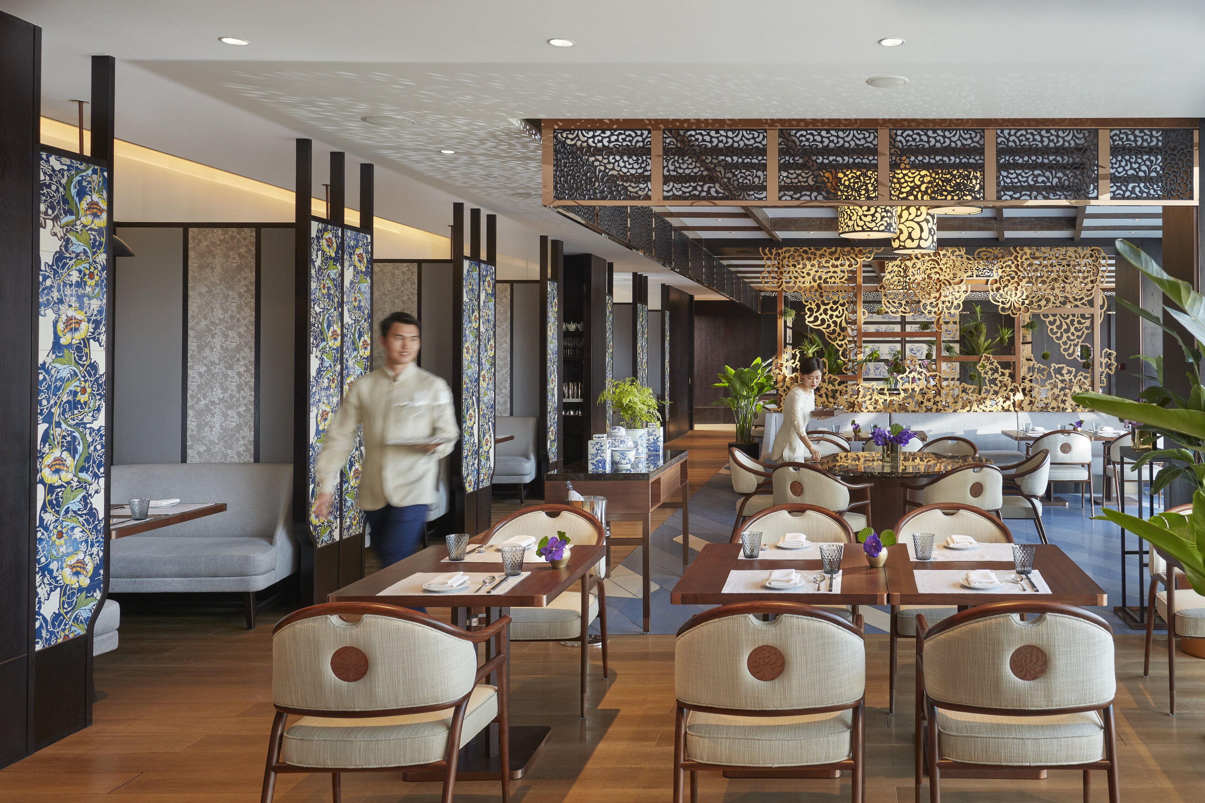 Een restaurant met modern interieur, met een eetgedeelte met tafels en stoelen, een decoratieve wand met ingewikkelde patronen, en een trap naar een hoger niveau.