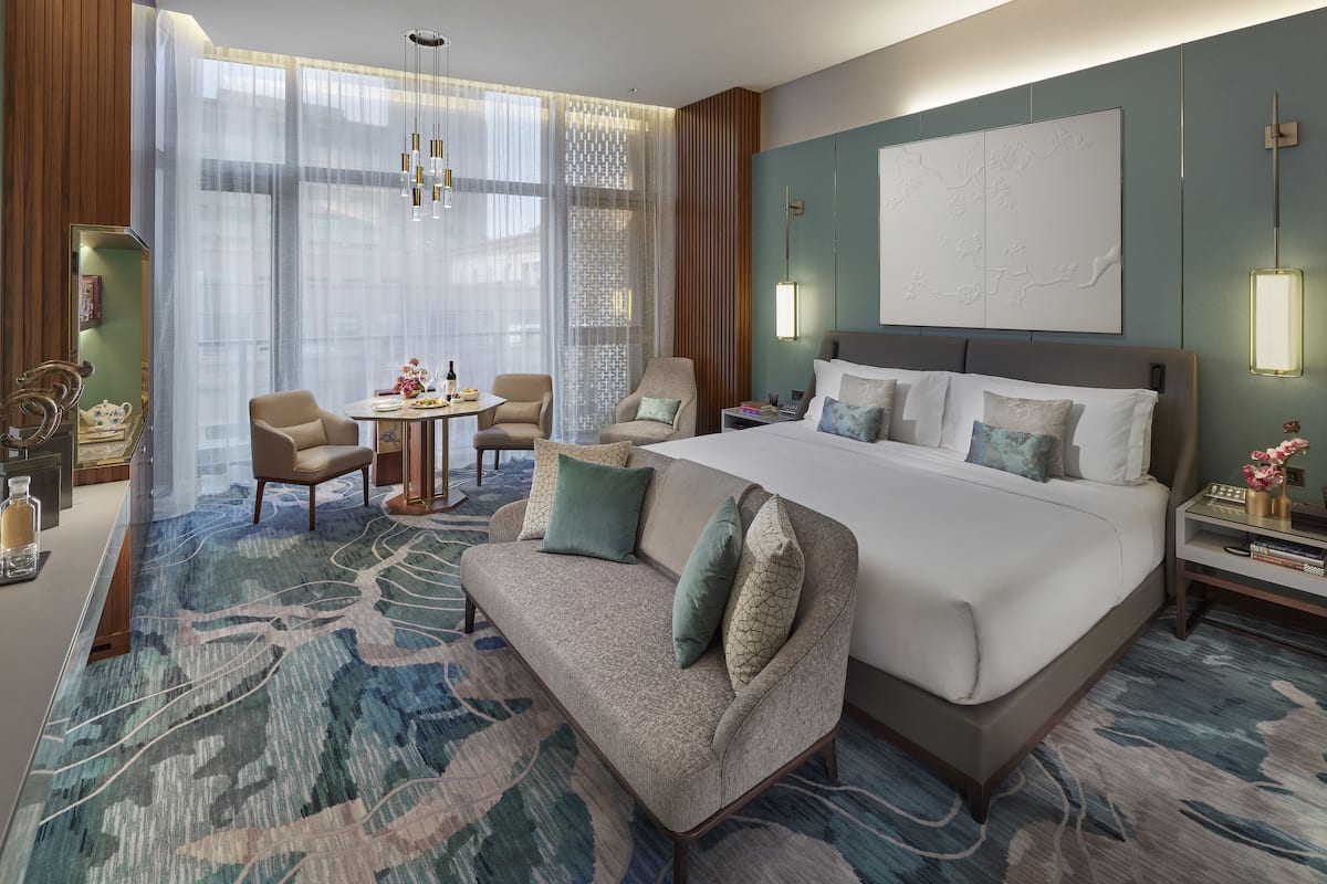 Une chambre d’hôtel avec un grand lit, un canapé, une table à manger et une chaise. Il y a une grande fenêtre avec des rideaux légers, un lustre et une œuvre d’art accrochée au mur.