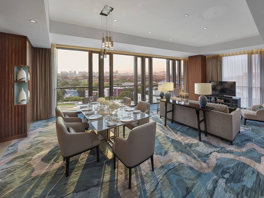 A spacious hotel room with a dining area, a living area with a TV, and large windows offering a city view.