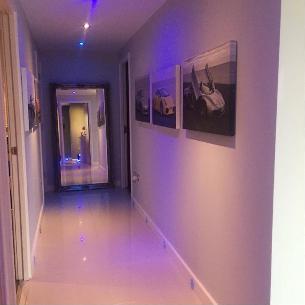Un couloir au sol et aux murs blancs, avec une porte au fond et trois tableaux encadrés accrochés au mur.