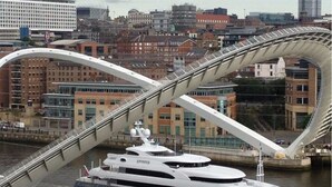 Eine große Yacht liegt unter einer modernen Brücke, im Hintergrund eine Stadtansicht.