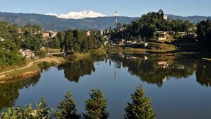 A serene lakeside view with a mountain backdrop, a bridge, and scattered buildings. - The Park Hotel (Kurseong)