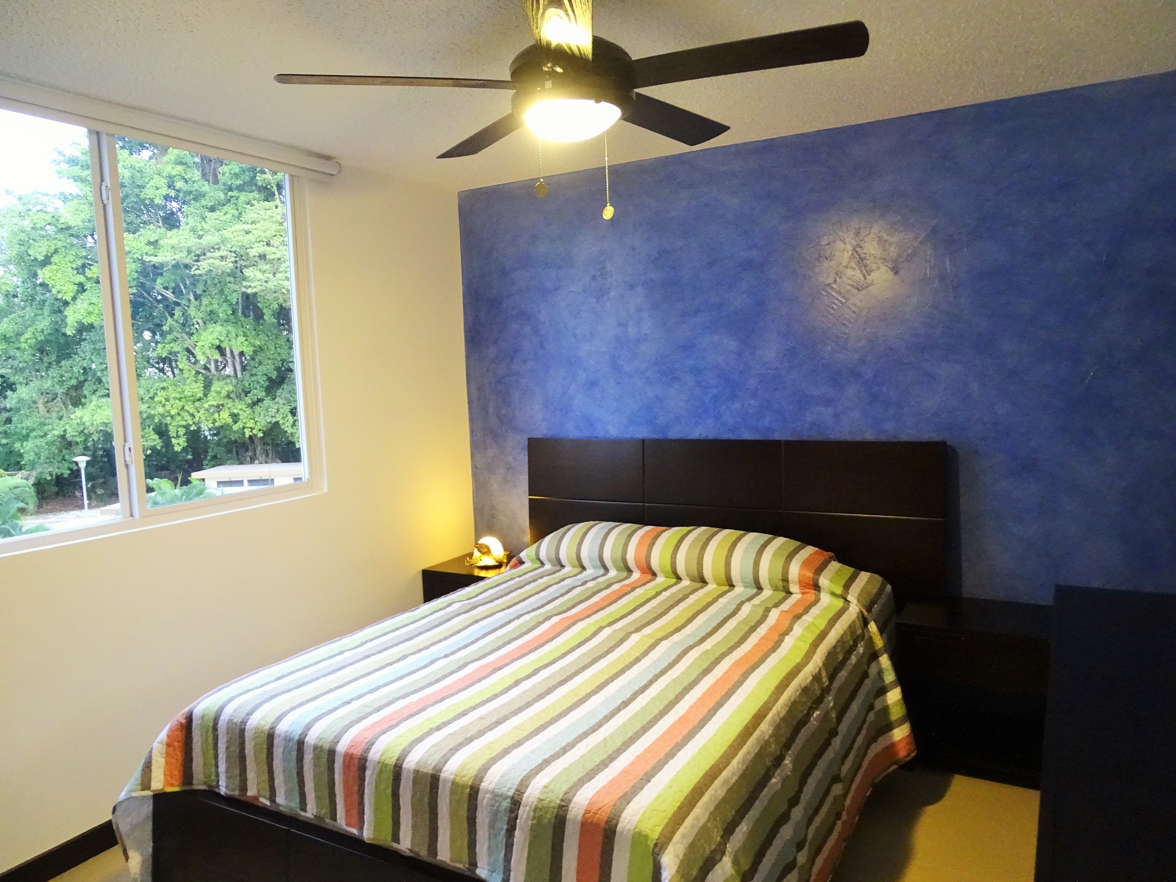 A bedroom with a bed, a nightstand, a ceiling fan, a window, and a view of trees outside.