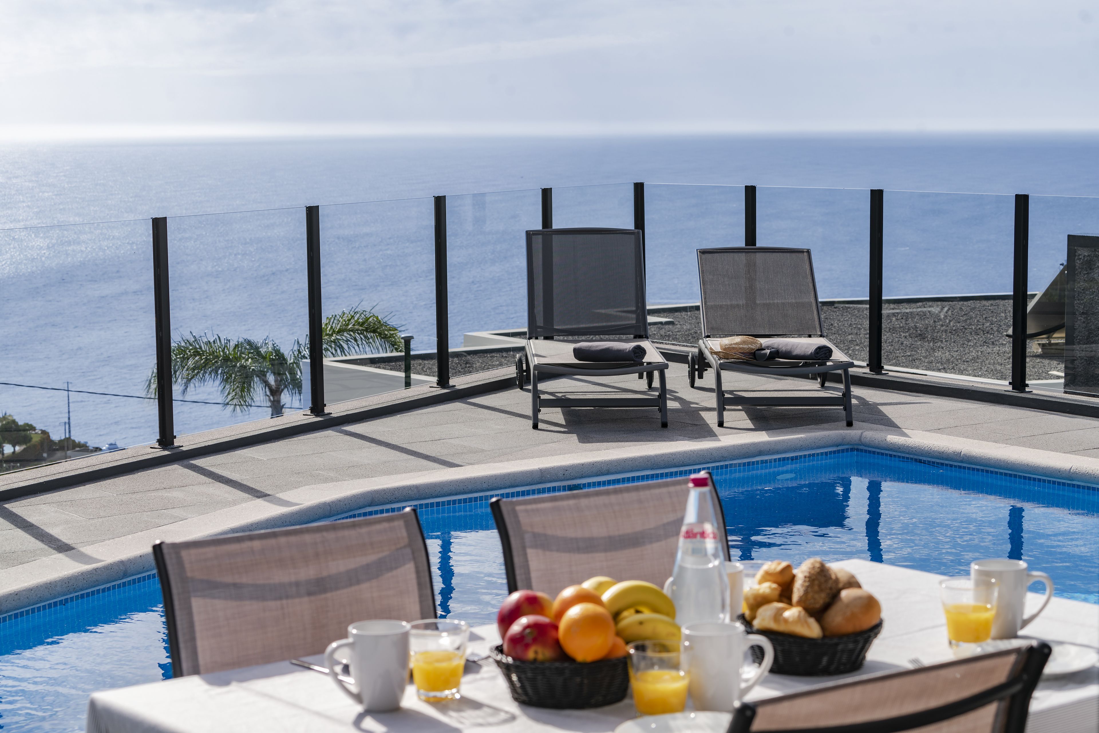 A poolside dining area with a view of the ocean, equipped with lounge chairs and a table set with breakfast items.