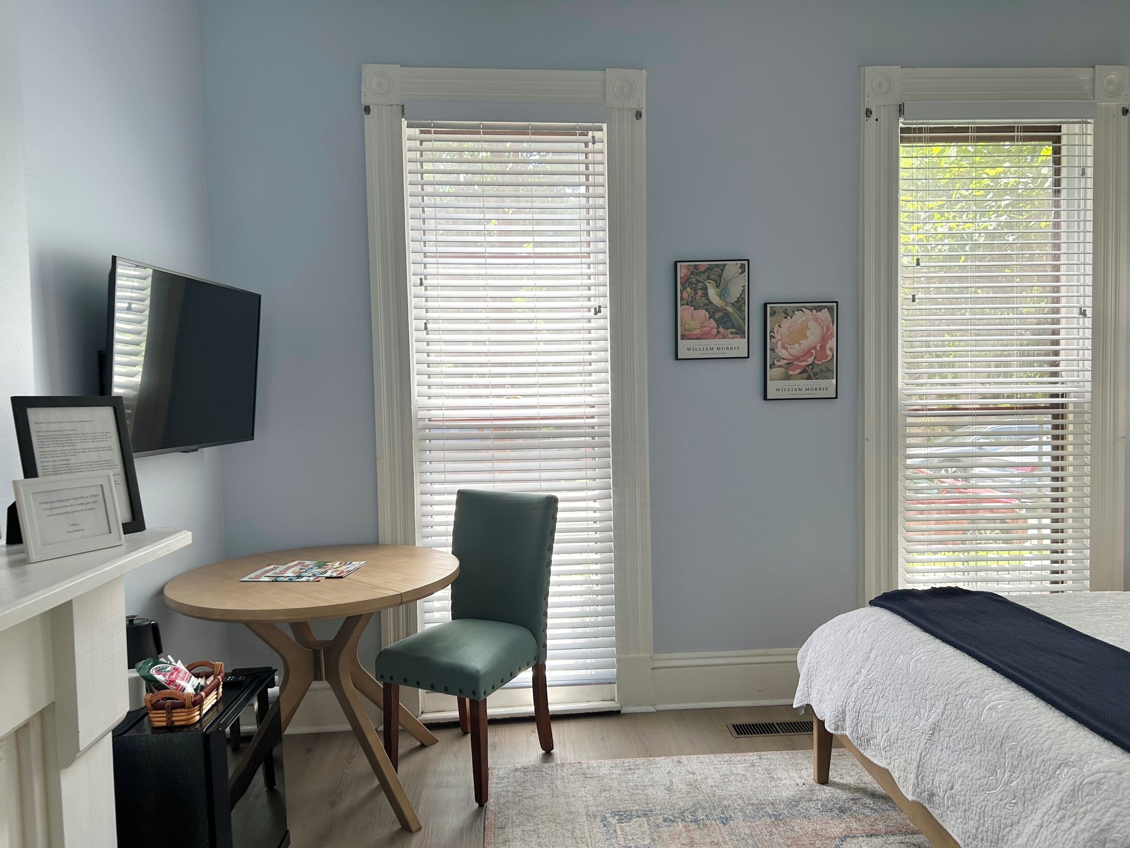 A bedroom with a bed, a desk with a chair, a television, and two windows with blinds.