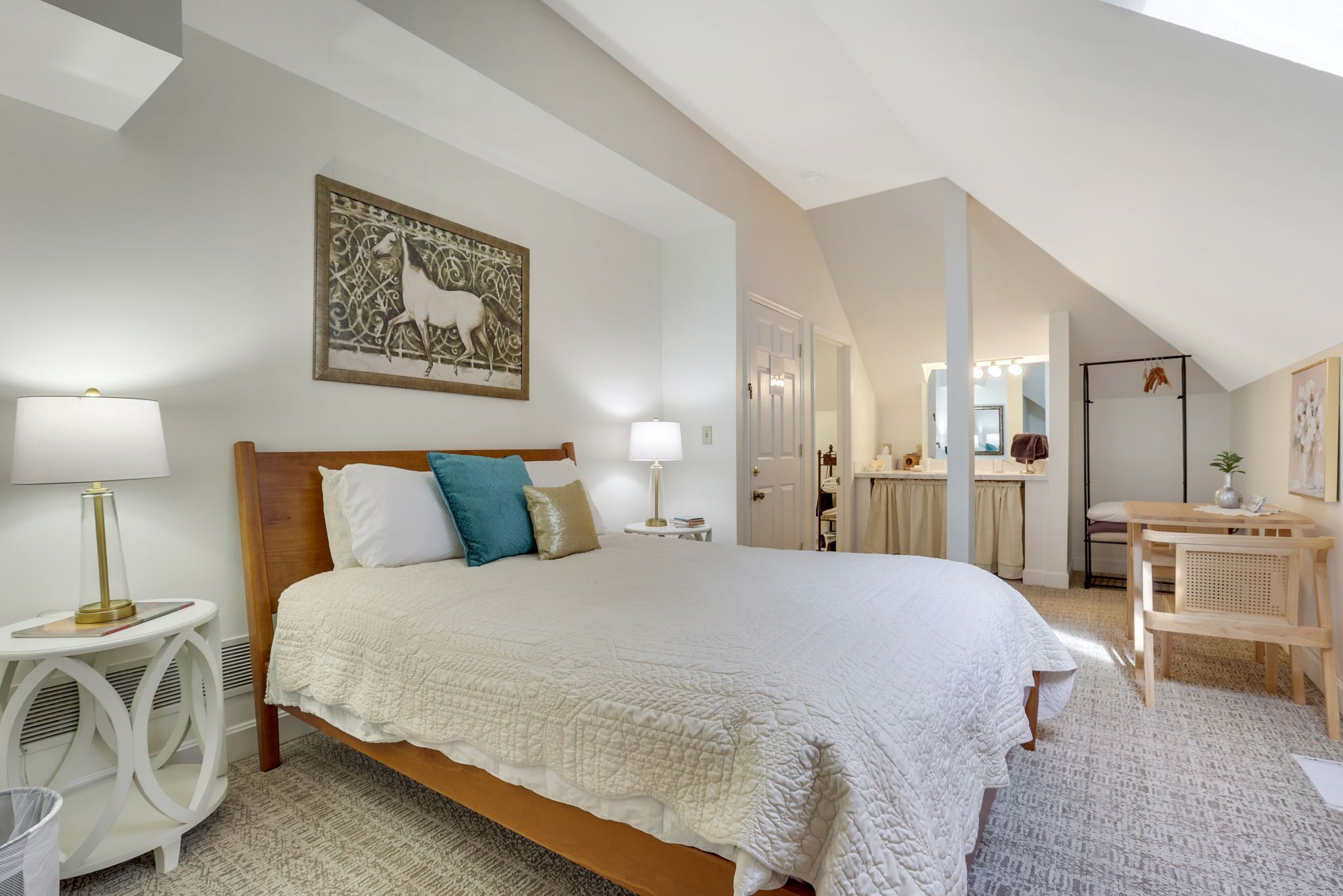 A spacious bedroom with a large bed, a bedside table with a lamp, a wall-mounted artwork, and a view into the adjacent bathroom.