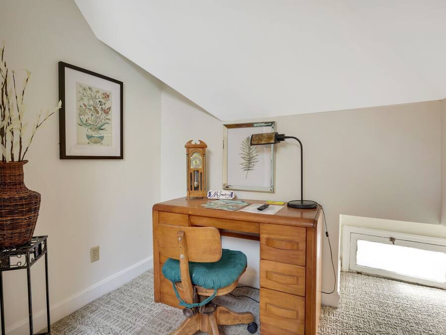 Un estudio con escritorio de madera, una silla, un jarrón, un grabado botánico enmarco y un reloj, además de una lámpara.