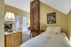 A bedroom with a brick chimney, a bed with white bedding, a desk with a lamp, and a framed floral artwork on the wall.