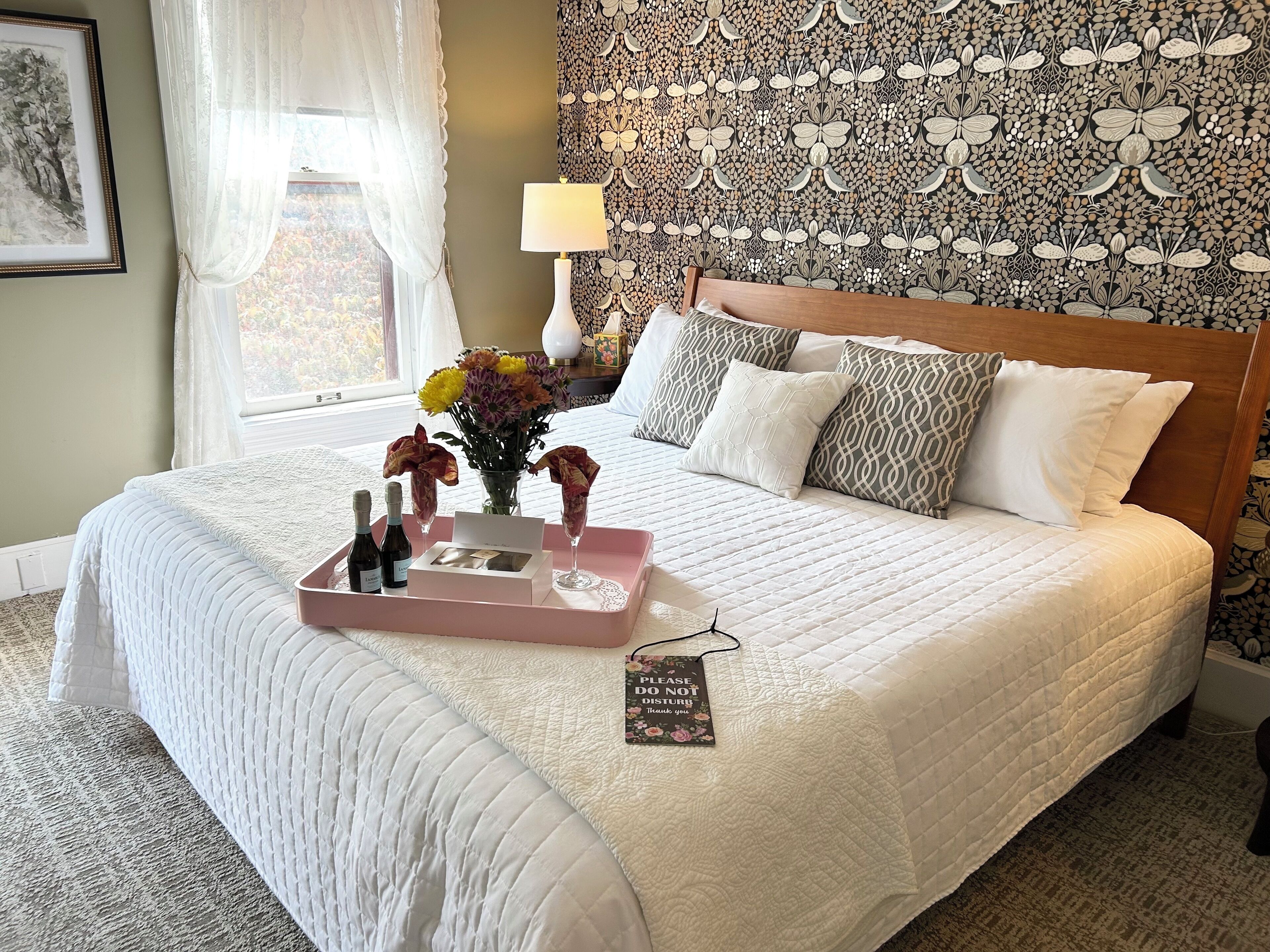 A neatly made bed with a tray of drinks and flowers, a bedside lamp, and a thank you card on the bed.