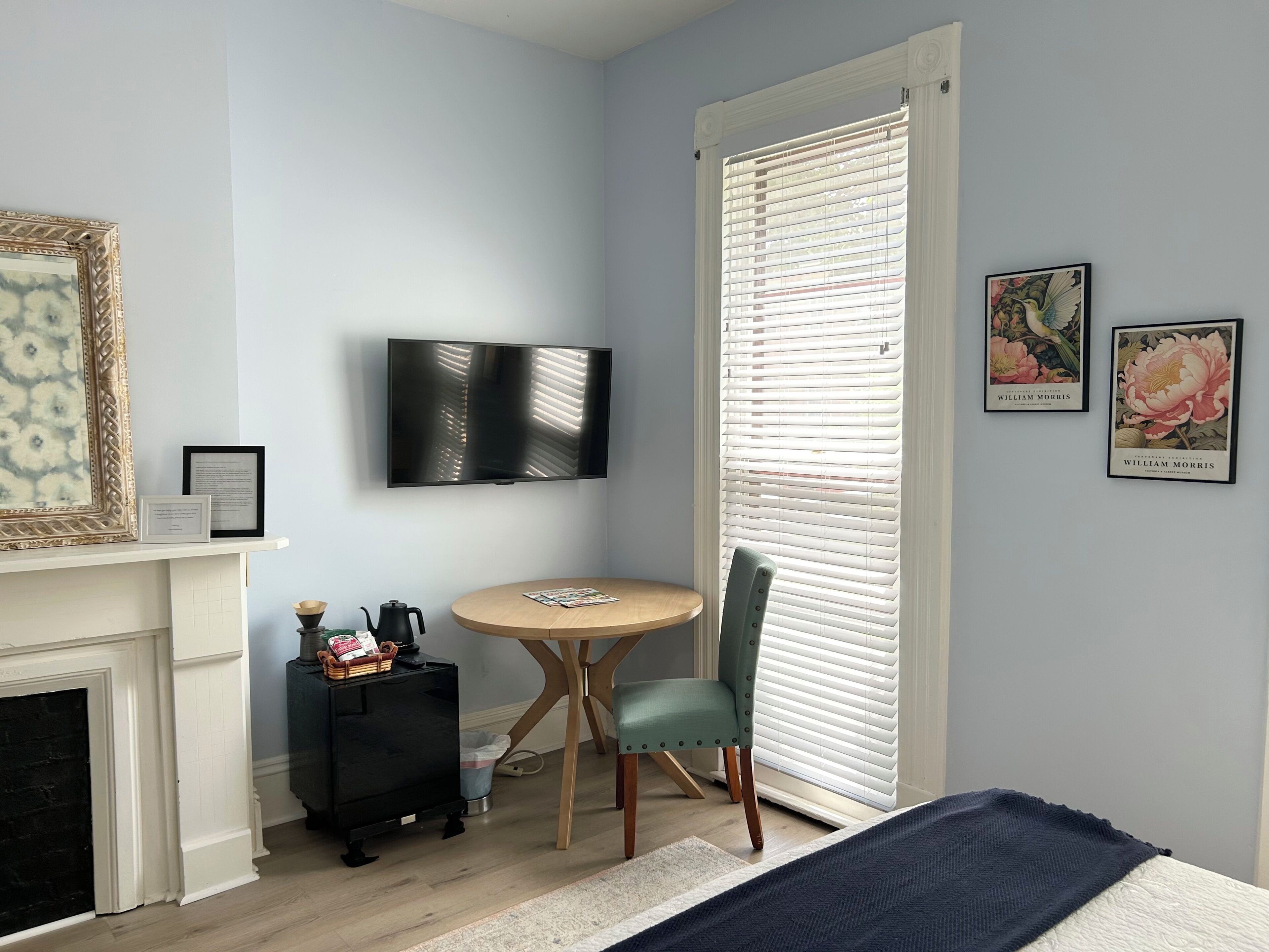 Une chambre à coucher comprenant un lit, un téléviseur fixé au mur, une cheminée, une table à manger avec une chaise, et une fenêtre avec des stores.