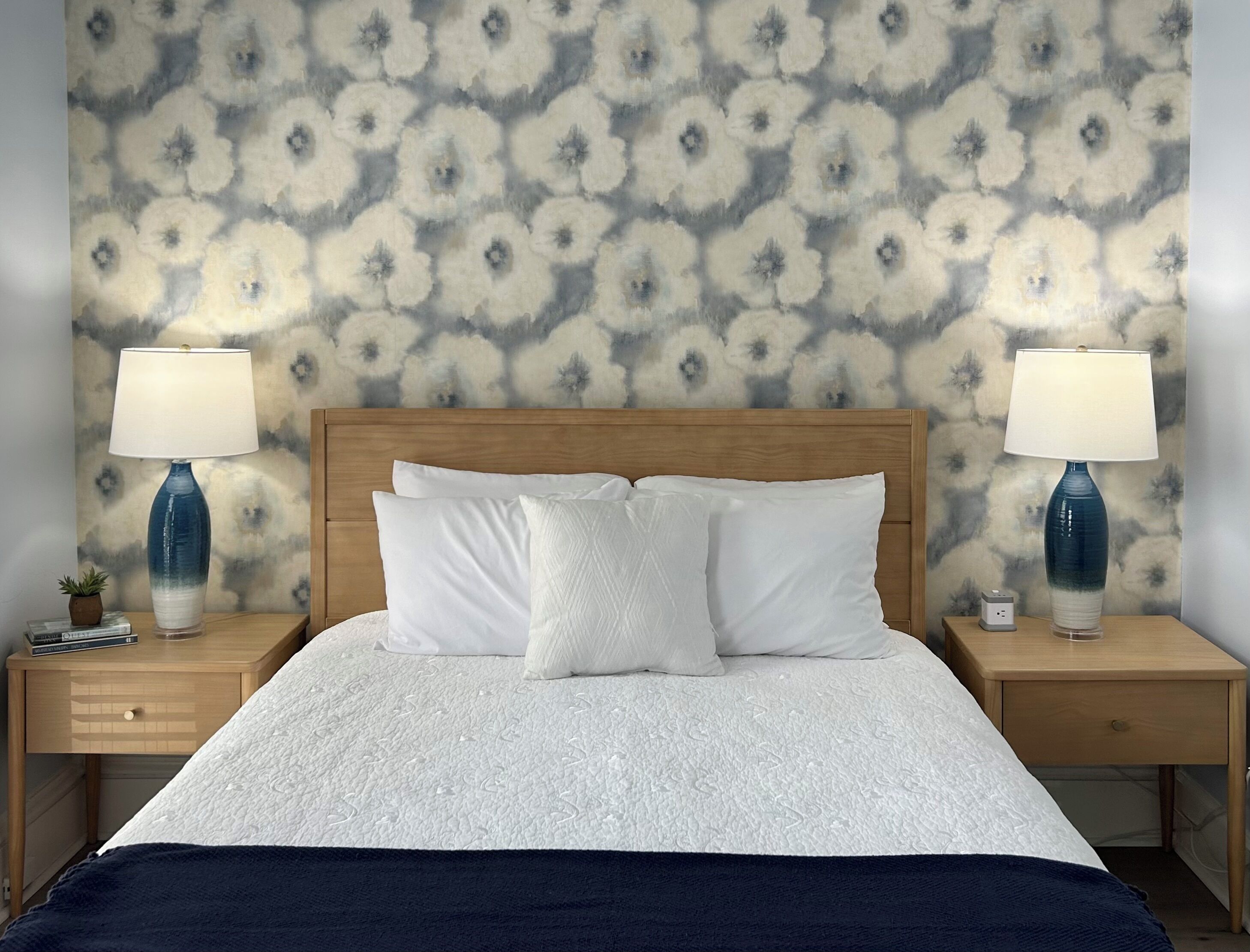 A neatly made bed with white bedding, two bedside tables with lamps, and a patterned wall behind.