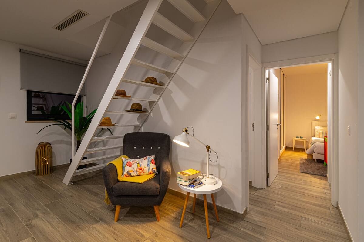 A modern living room with a staircase, a comfortable armchair, a small round table with books and a lamp, and a plant.