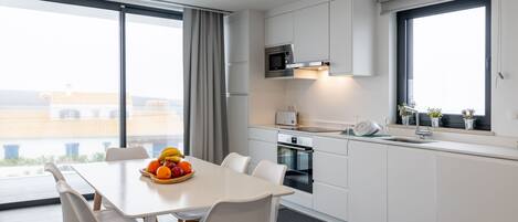 A modern kitchen with white cabinetry, a microwave, and a toaster. There is a dining table with chairs and a fruit bowl.