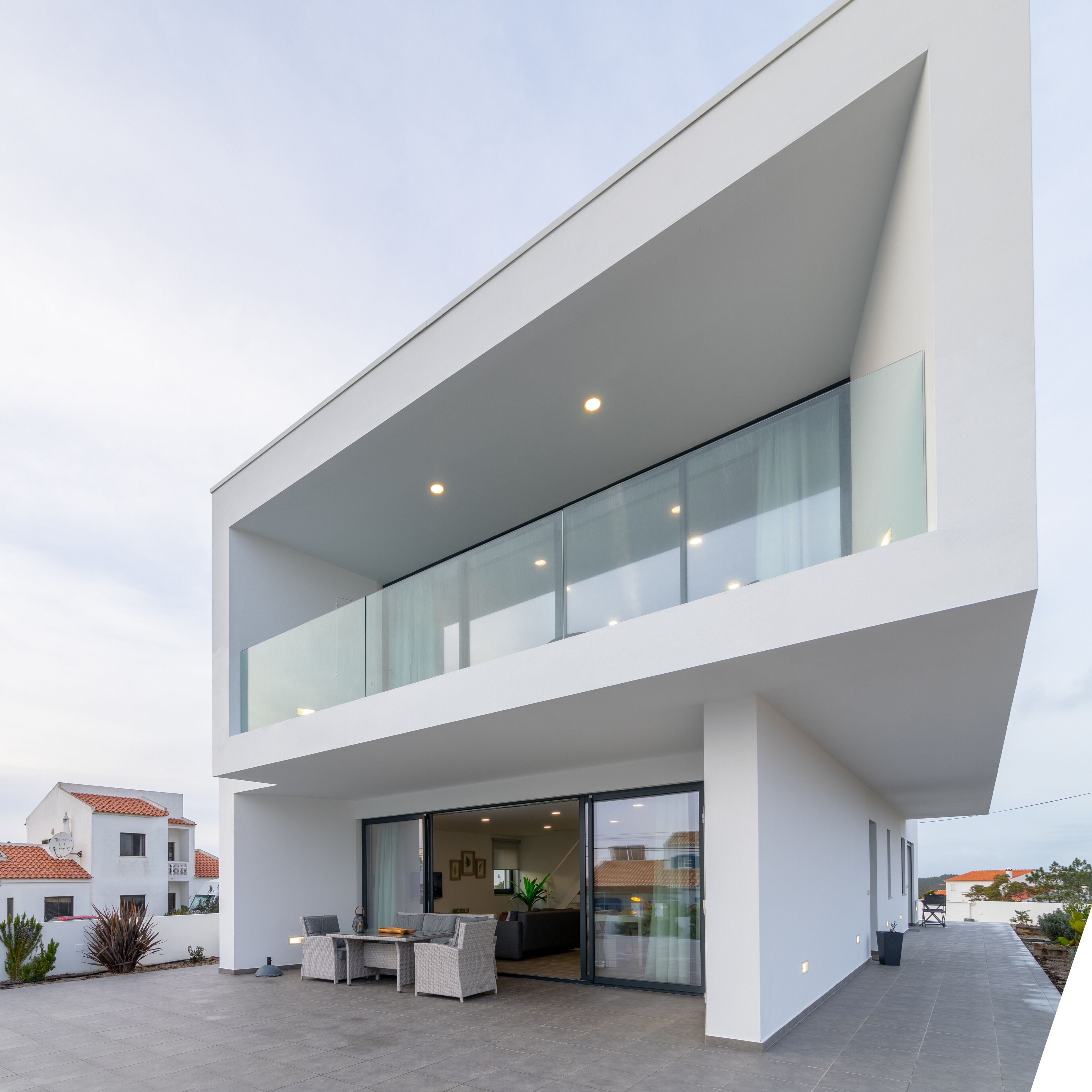 A modern two-story house with a flat roof, large glass windows, and a patio area with outdoor furniture.