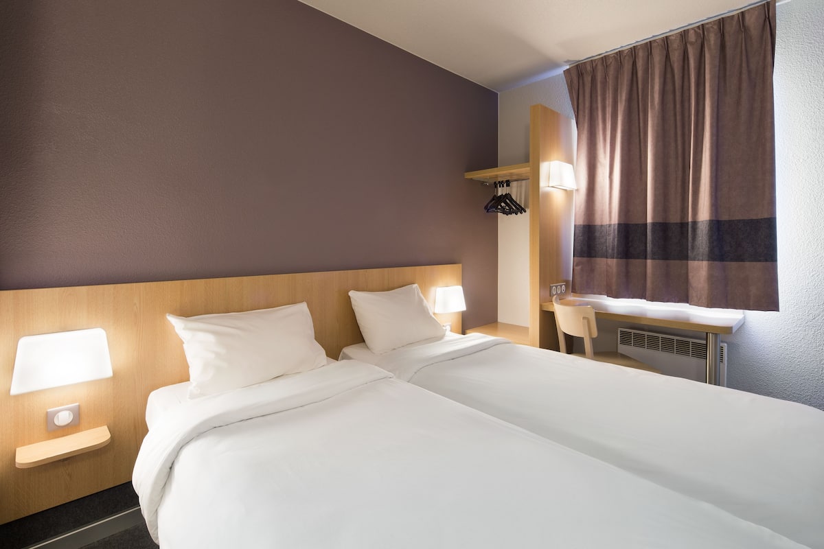 A hotel room with two beds, a desk, a chair, a window with curtains, and a wall-mounted lamp.
