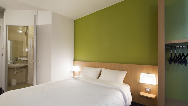 A hotel room with a bed, a nightstand with a lamp, a green accent wall, and a bathroom visible through an open door.
