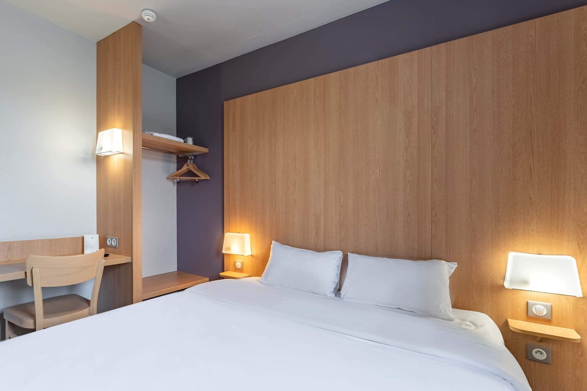 A hotel room with a large bed, bedside lamps, a wooden desk, and a hanging coat rack.