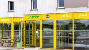 A hotel entrance with glass doors, a sign reading "Entrée," and a small outdoor seating area with tables and chairs.