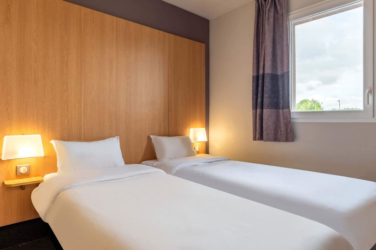 A hotel room with two single beds, wooden headboard, bedside lamps, and a window with curtains.