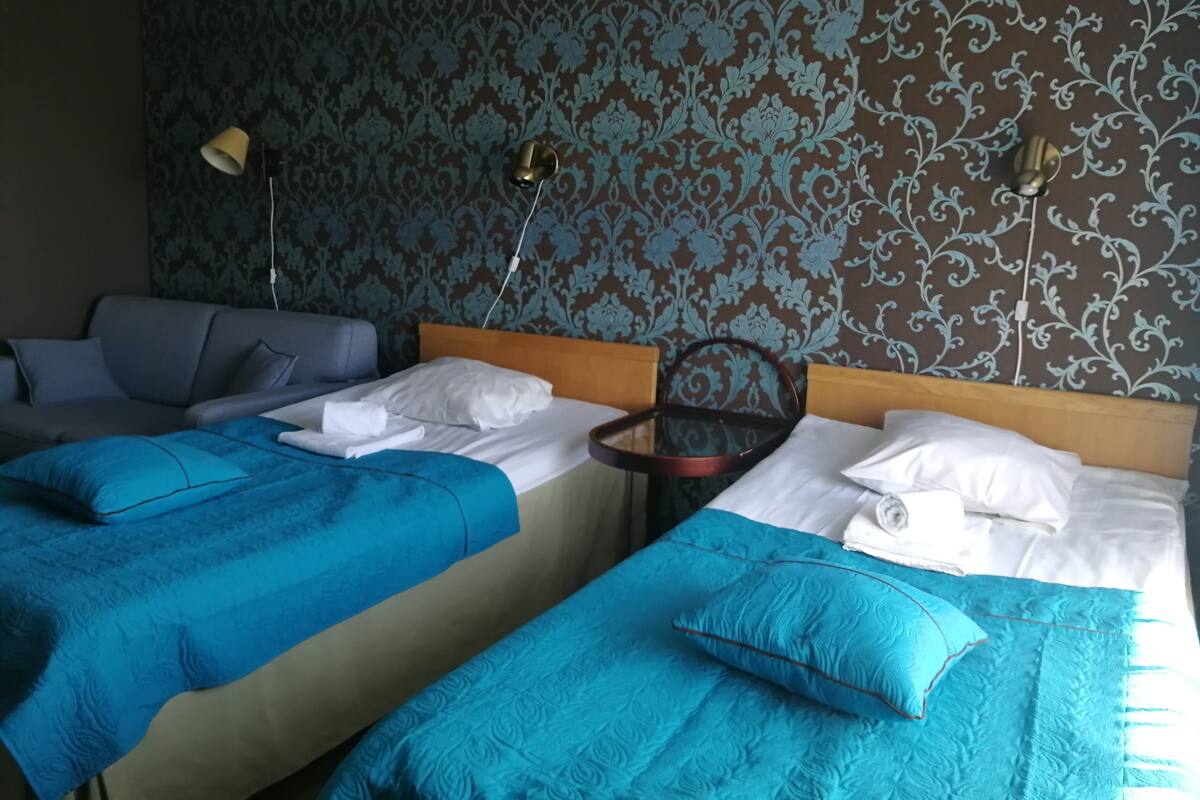 A hotel room with two beds, each with a blue bedspread and white pillows. There is a wooden headboard, a bedside table with a lamp, and a patterned wallpaper.