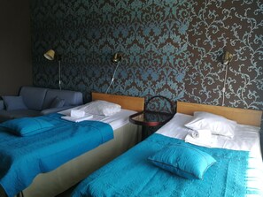 A hotel room with two beds, each with a blue bedspread and white pillows. There is a wooden headboard, a bedside table with a lamp, and a patterned wallpaper. - Hotel Villa Vanessa (Pyhtaa)