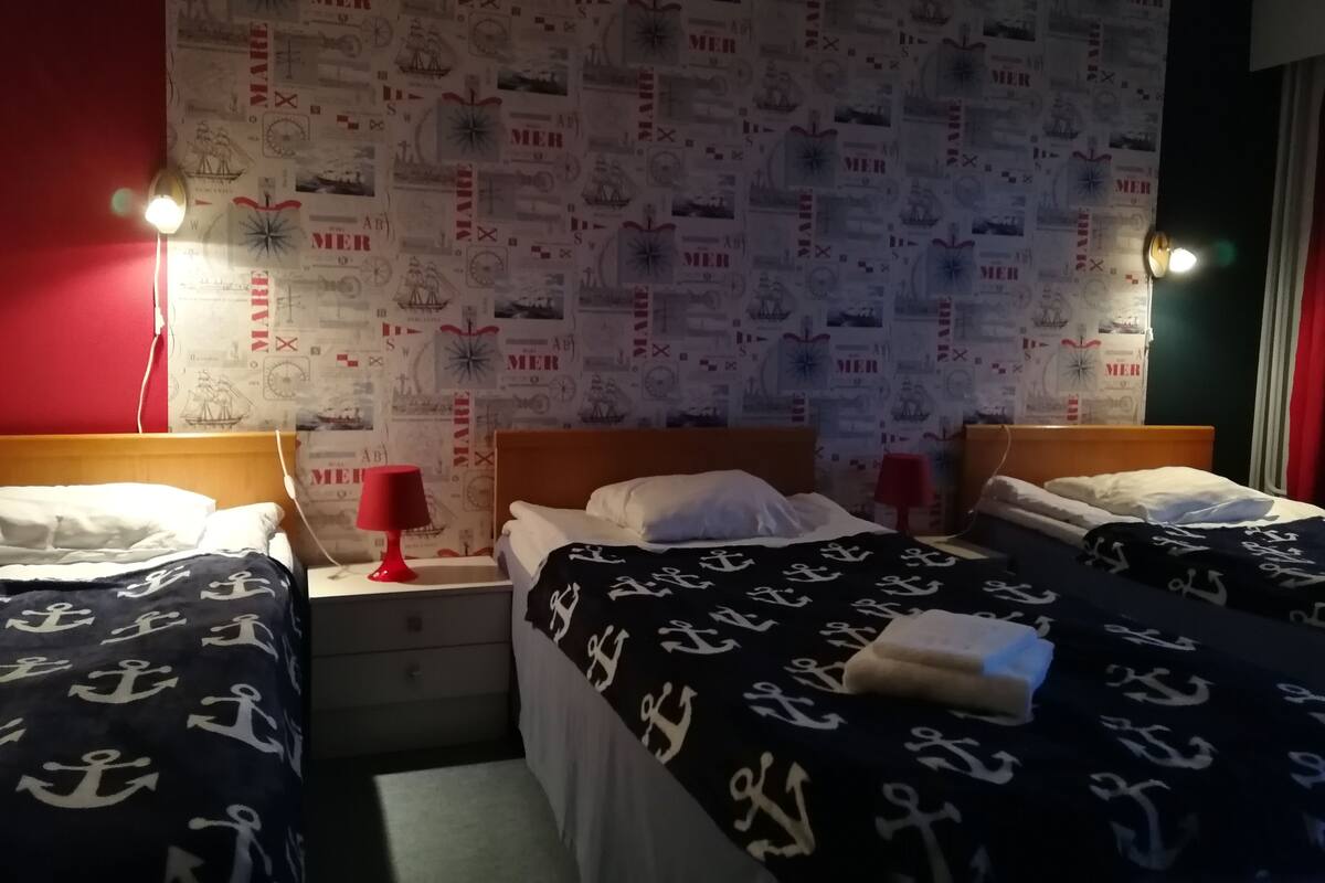 A hotel room with two beds, each with a navy blue blanket featuring white anchors. The walls are adorned with a maritime-themed wallpaper. There are two bedside tables with lamps, and the room has a red accent wall.