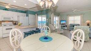 A spacious living area with a dining table, white chairs, a kitchen with white cabinets, a ceiling fan, and a view of the ocean through large windows.