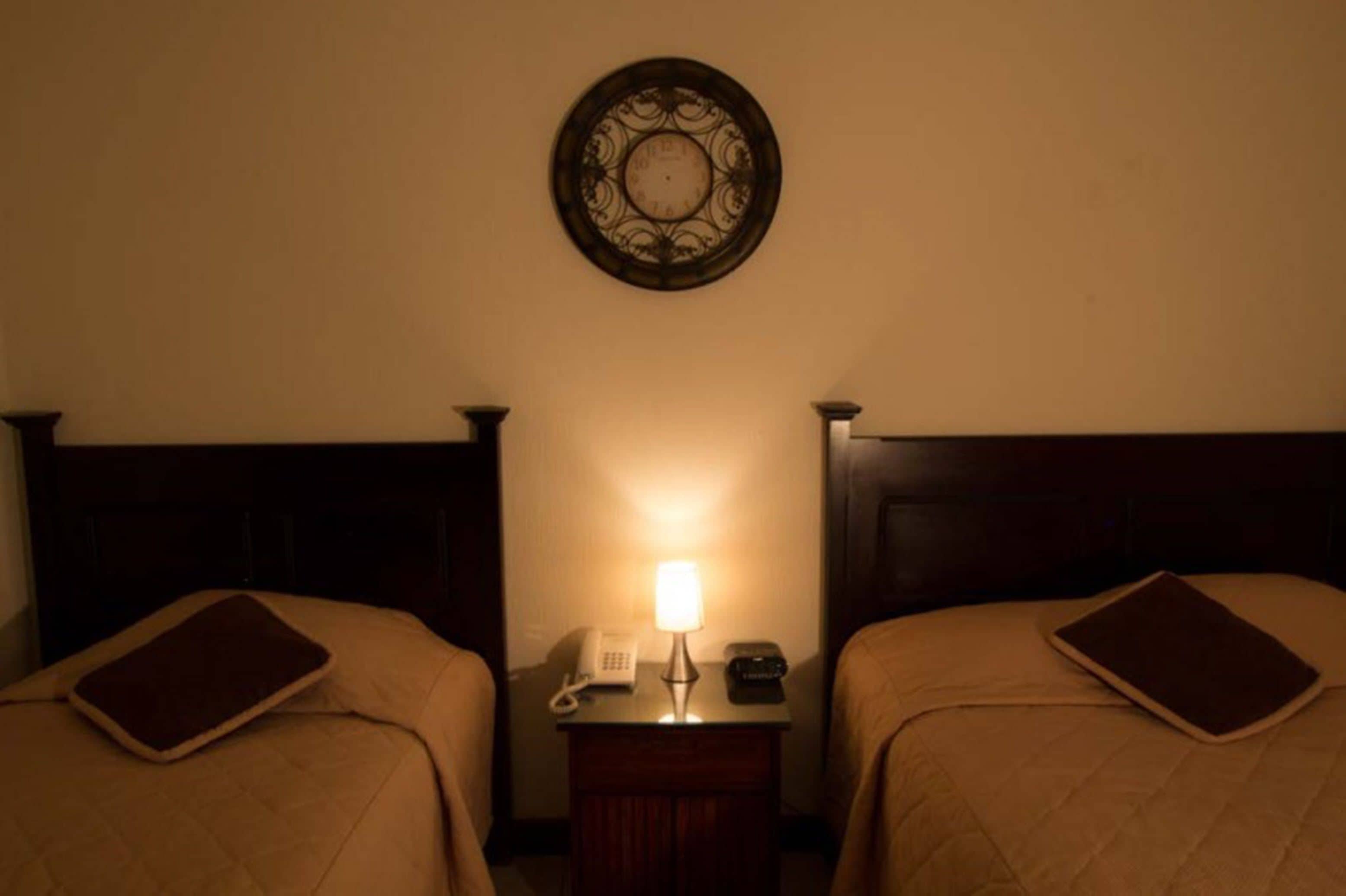 Ein Hotelzimmer mit zwei Betten, einem Nachttisch mit Lampe, einem Telefon und einer Wanduhr.
