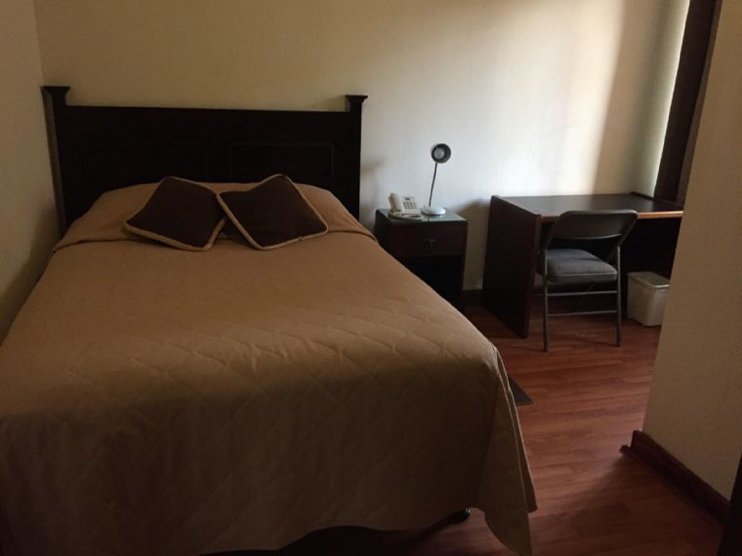 A neatly made bed with a dark headboard, a bedside table with a lamp, and a chair in a room with wooden flooring.