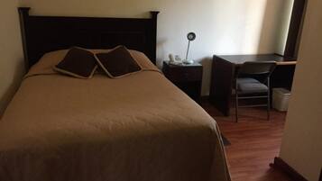 A neatly made bed with a dark headboard, a bedside table with a lamp, and a chair in a room with wooden flooring.