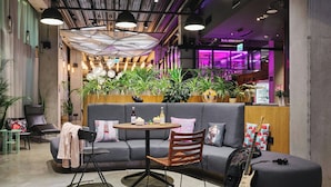 Un área de descanso moderna con una mesa redonda, un sofá y un sillón. Se observan plantas, una guitarra y un refrigerador al fondo.