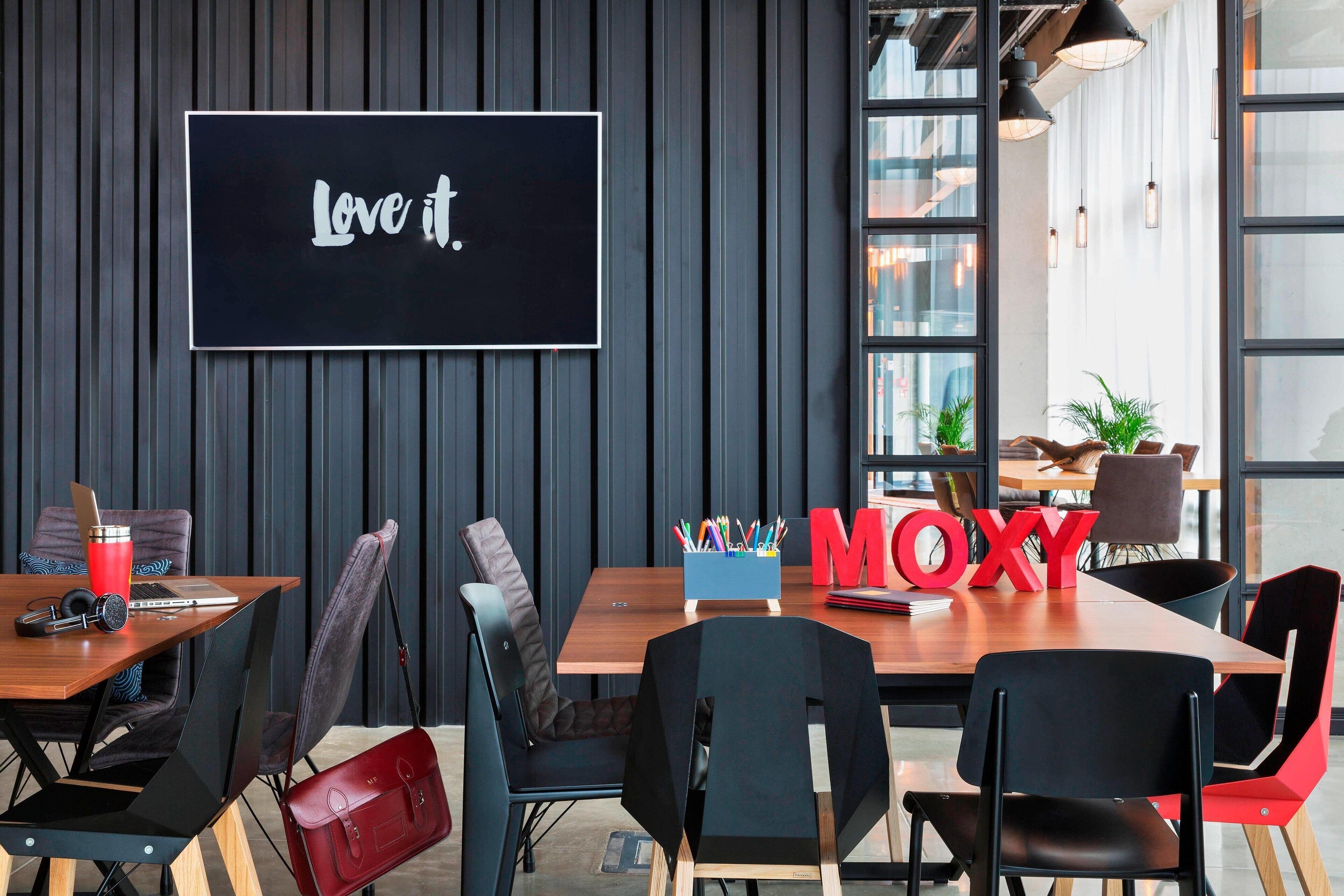 A modern meeting room with a large screen displaying "Love it.", a wooden table with chairs, and a "MOXY" sign.