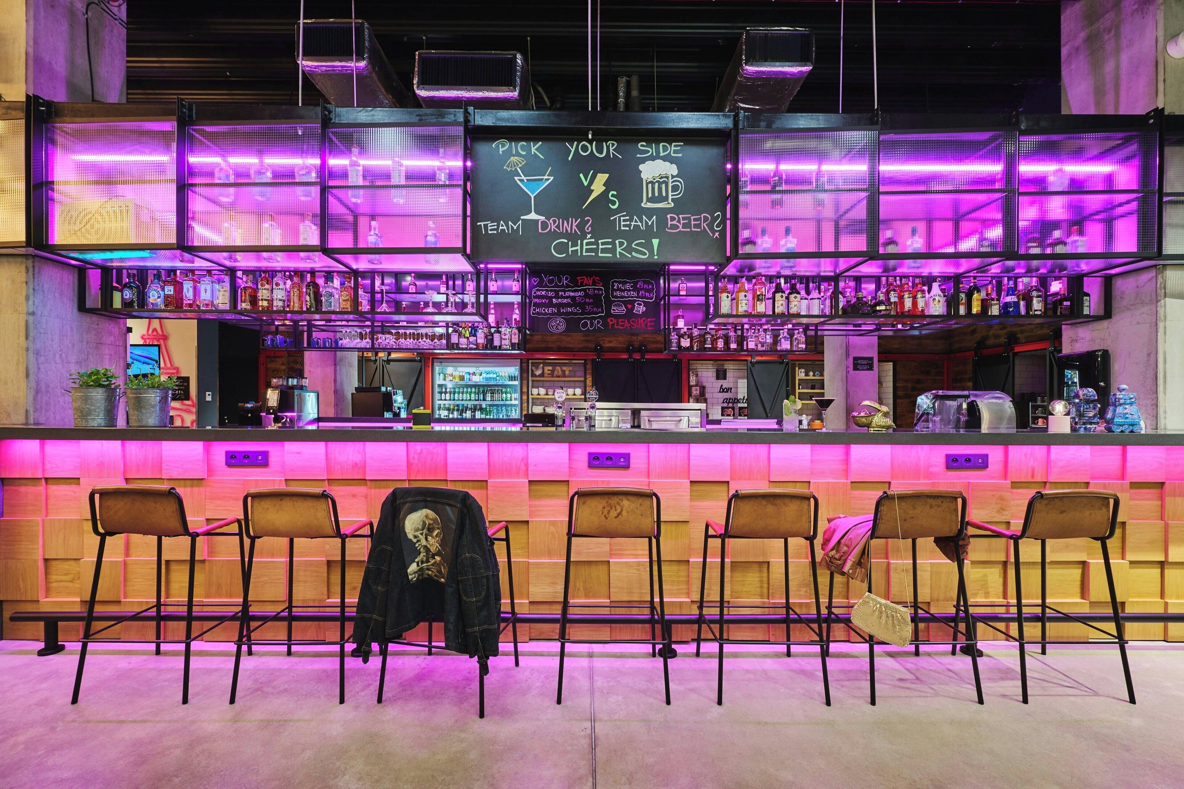A bar area with a neon sign, bar stools, and a variety of bottles on shelves.