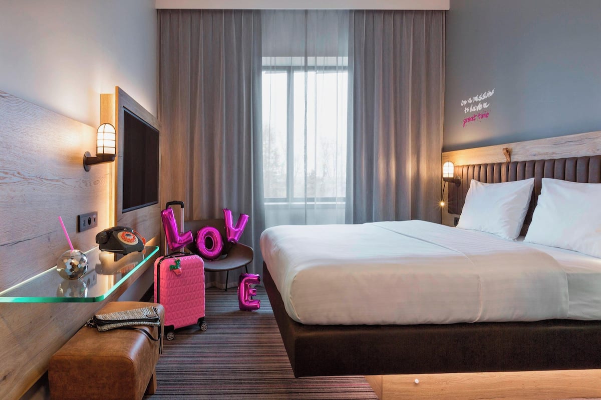 Una habitación de hotel con cama, mesita de noche, lámpara, televisor y ventana con cortinas.