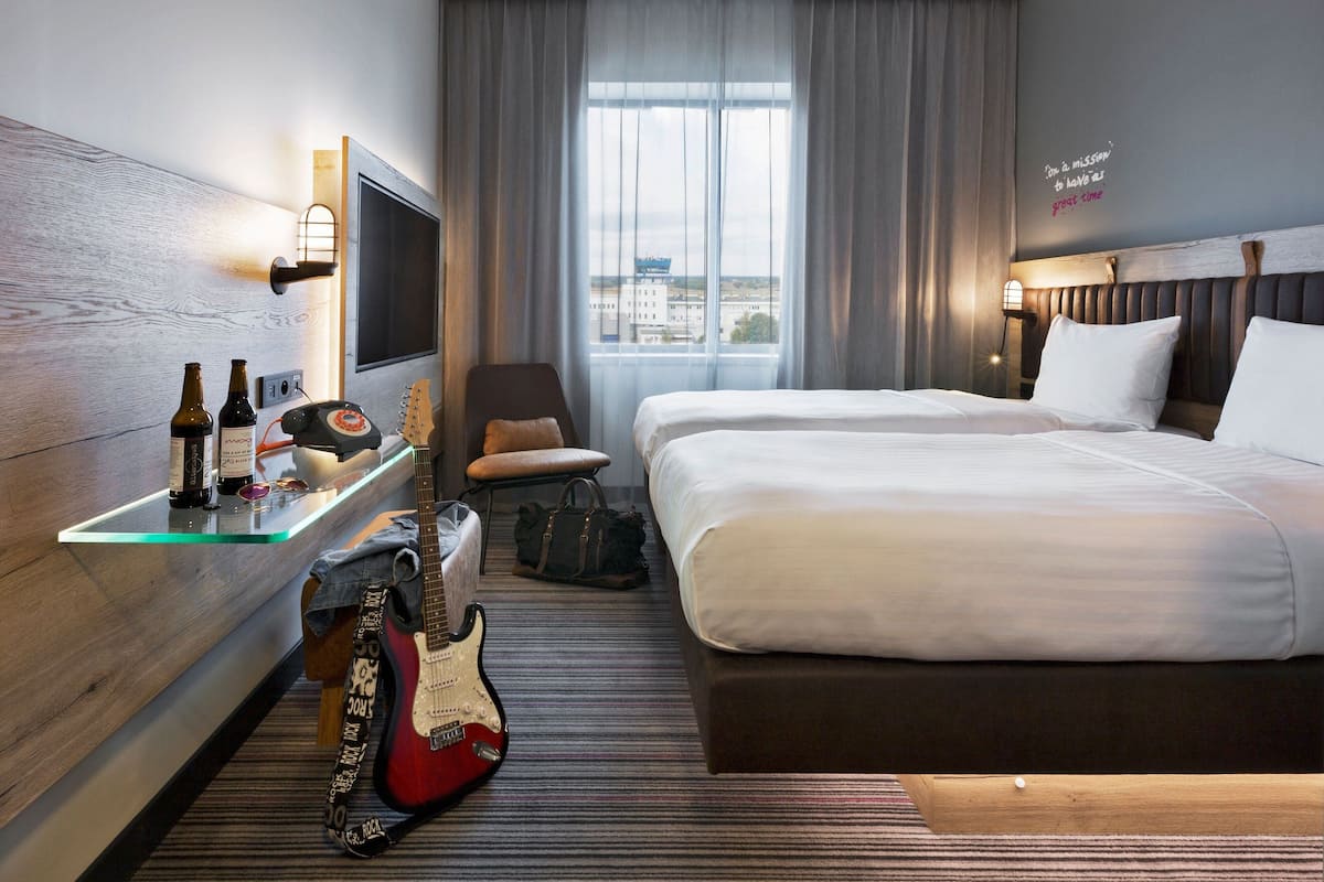 Una habitación de hotel con cama, escritorio, guitarra, silla y ventana con vista a la ciudad.