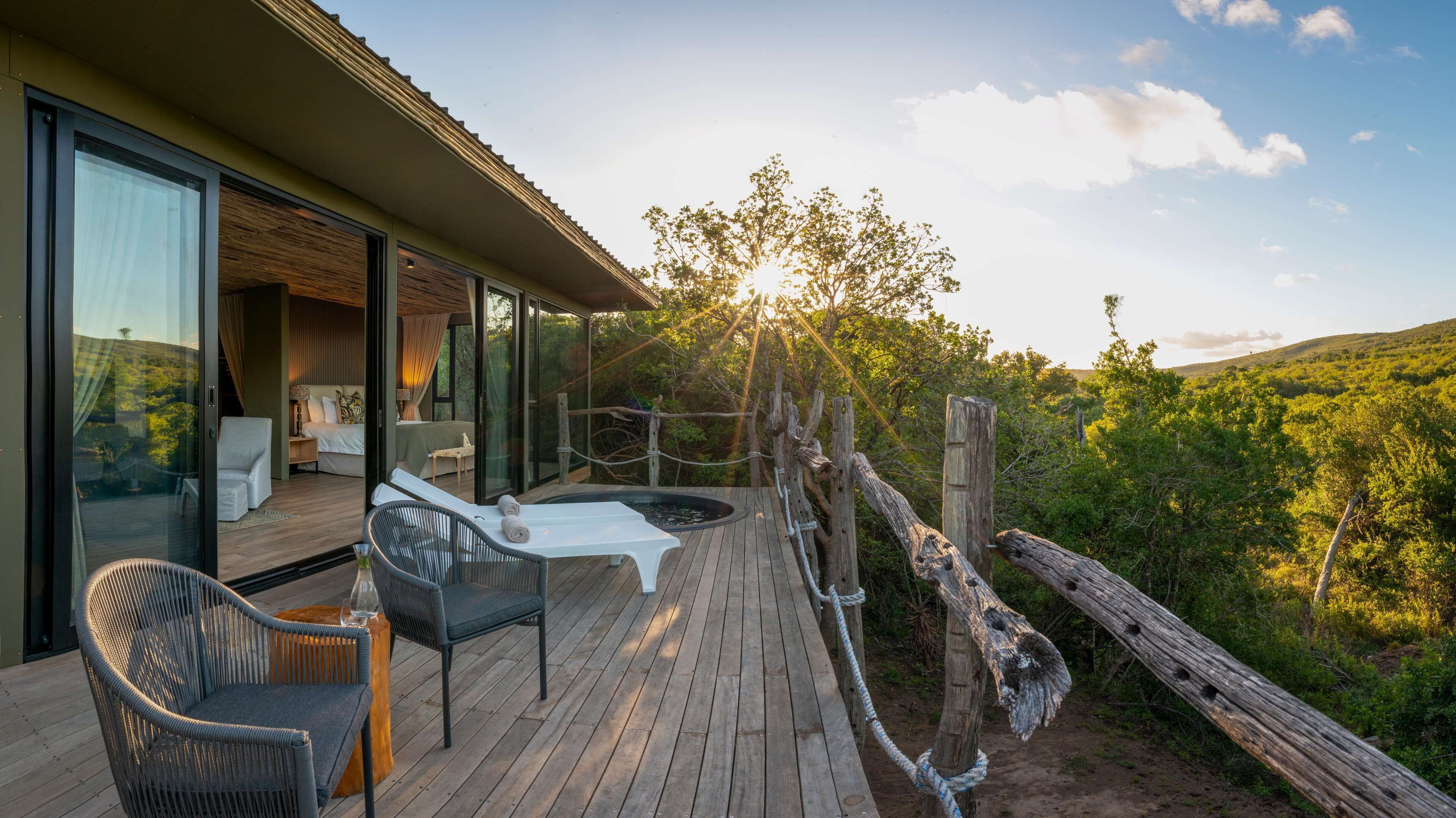 Een houten terras met buitenmeubilair, een jacuzzi en een slaapkamer met een groot raam met uitzicht op een bebost gebied.