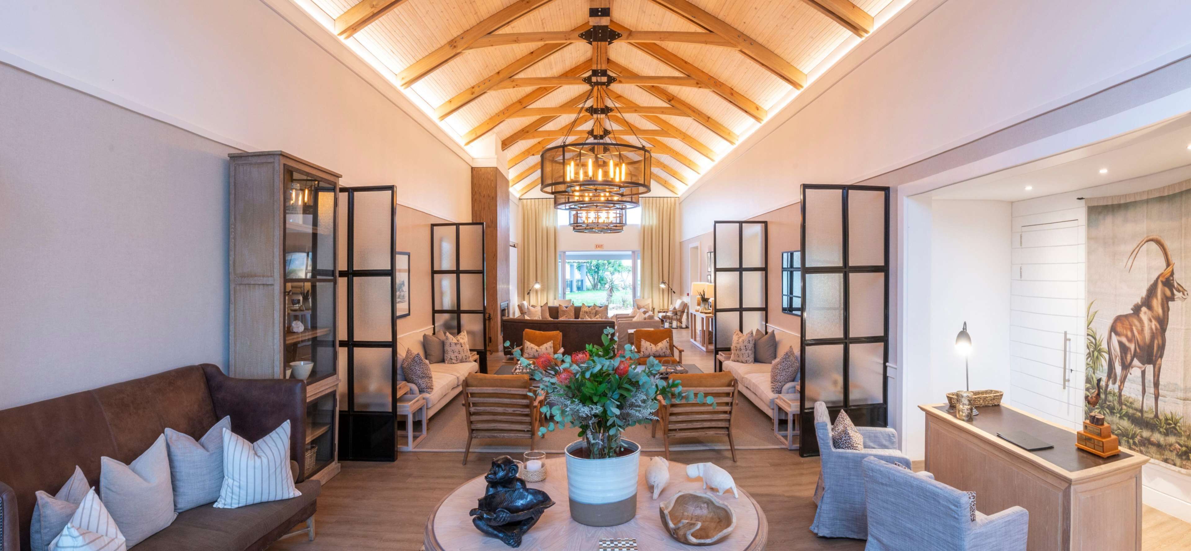 A spacious living room with a high ceiling, wooden beams, and a central chandelier. There are multiple seating areas with sofas and chairs, and a large window providing natural light.