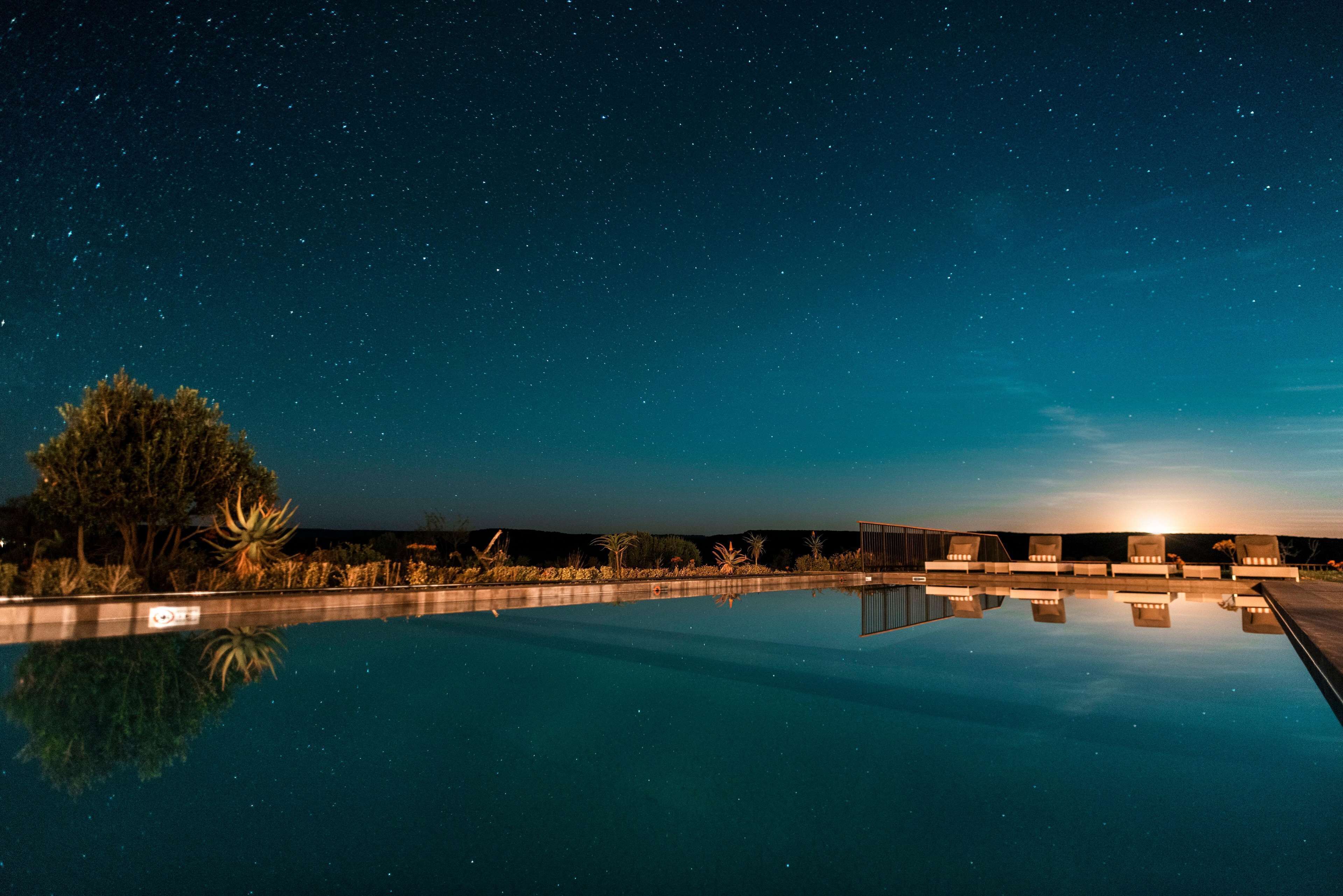 A nighttime pool area with a clear view of the sky and stars, surrounded by trees and a row of chairs.