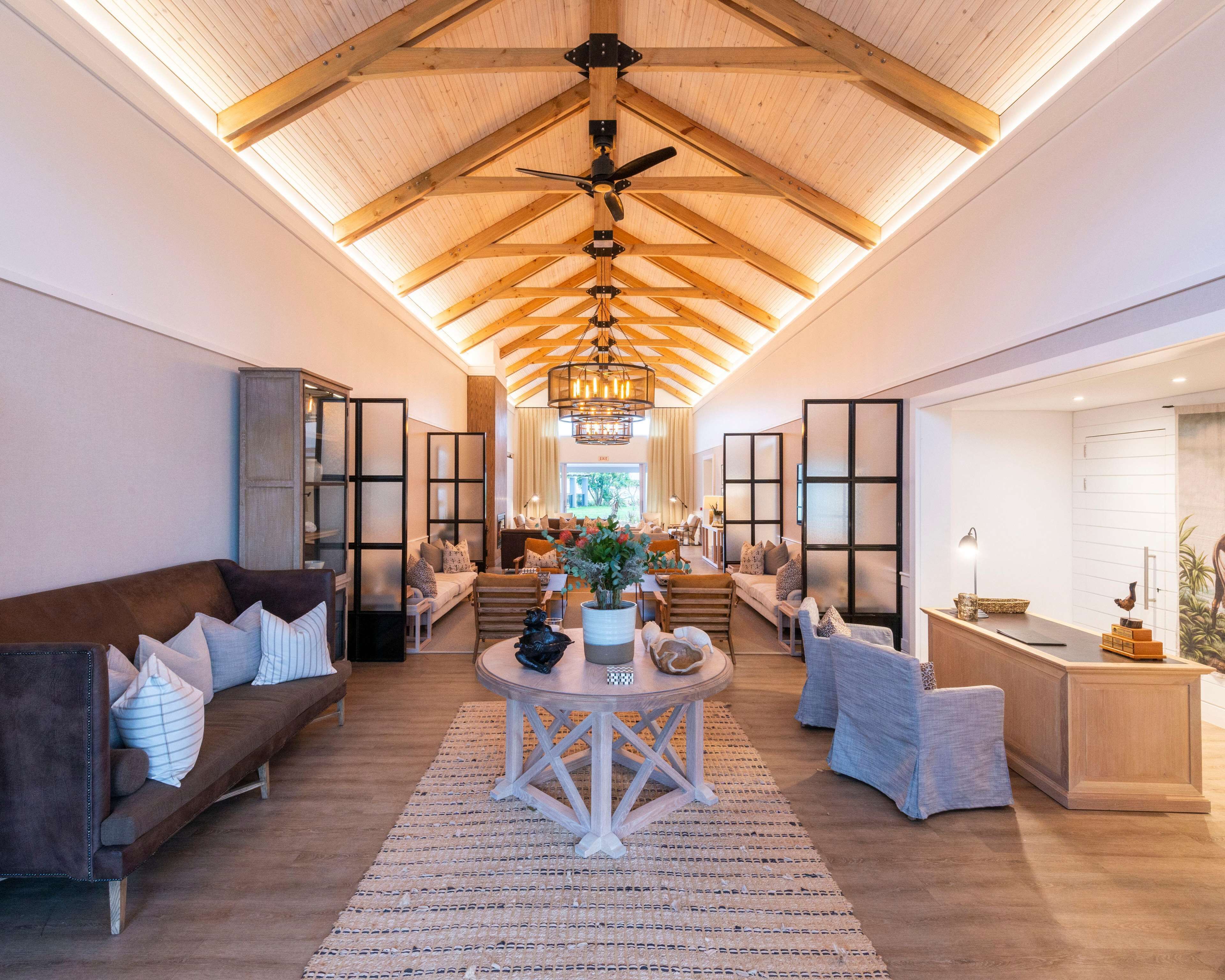 A spacious living area with a high ceiling, wooden beams, a central chandelier, and a view of the kitchen through a glass partition.