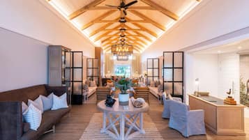 A spacious living area with a high ceiling, wooden beams, a central chandelier, and a view of the kitchen through a glass partition.