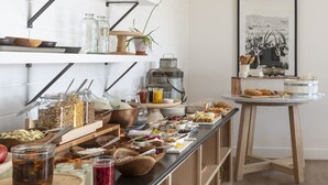 Disposição de buffet com diversos alimentos e acompanhamentos sobre uma mesa comprida, rodeada por prateleiras com utensílios de cozinha e uma moldura com imagem na parede.