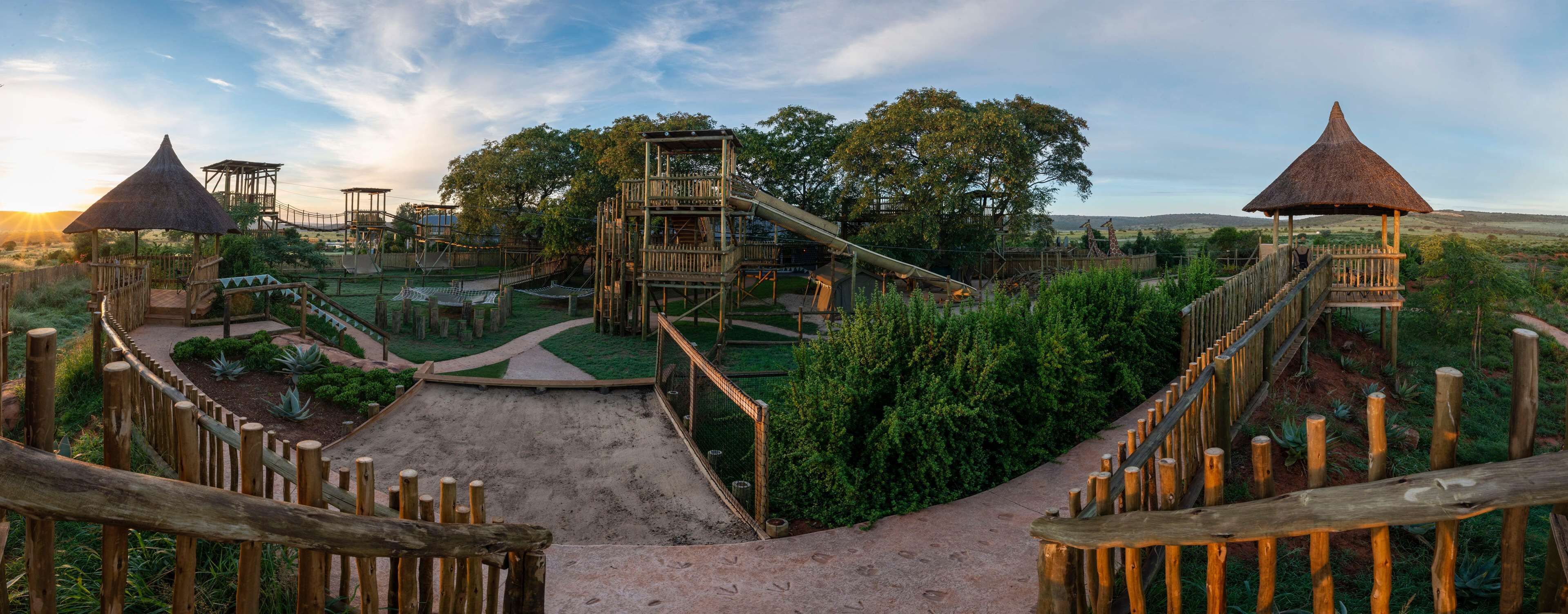Un terrain de jeux avec des structures et des toboggans en bois, entouré de verdure et sous un ciel dégagé.