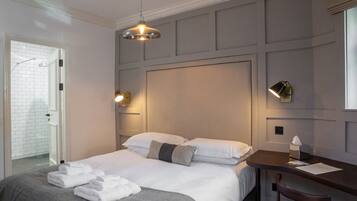 Une chambre d’hôtel avec un lit bien fait, un petit bureau et une chaise. On aperçoit l’espace de la douche à travers une porte ouverte.
