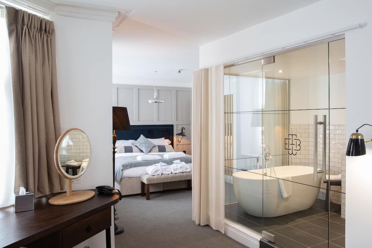 A hotel room with a bed, a desk with a mirror, a bedside lamp, a large window with curtains, a glass-enclosed shower area, and a bathtub.