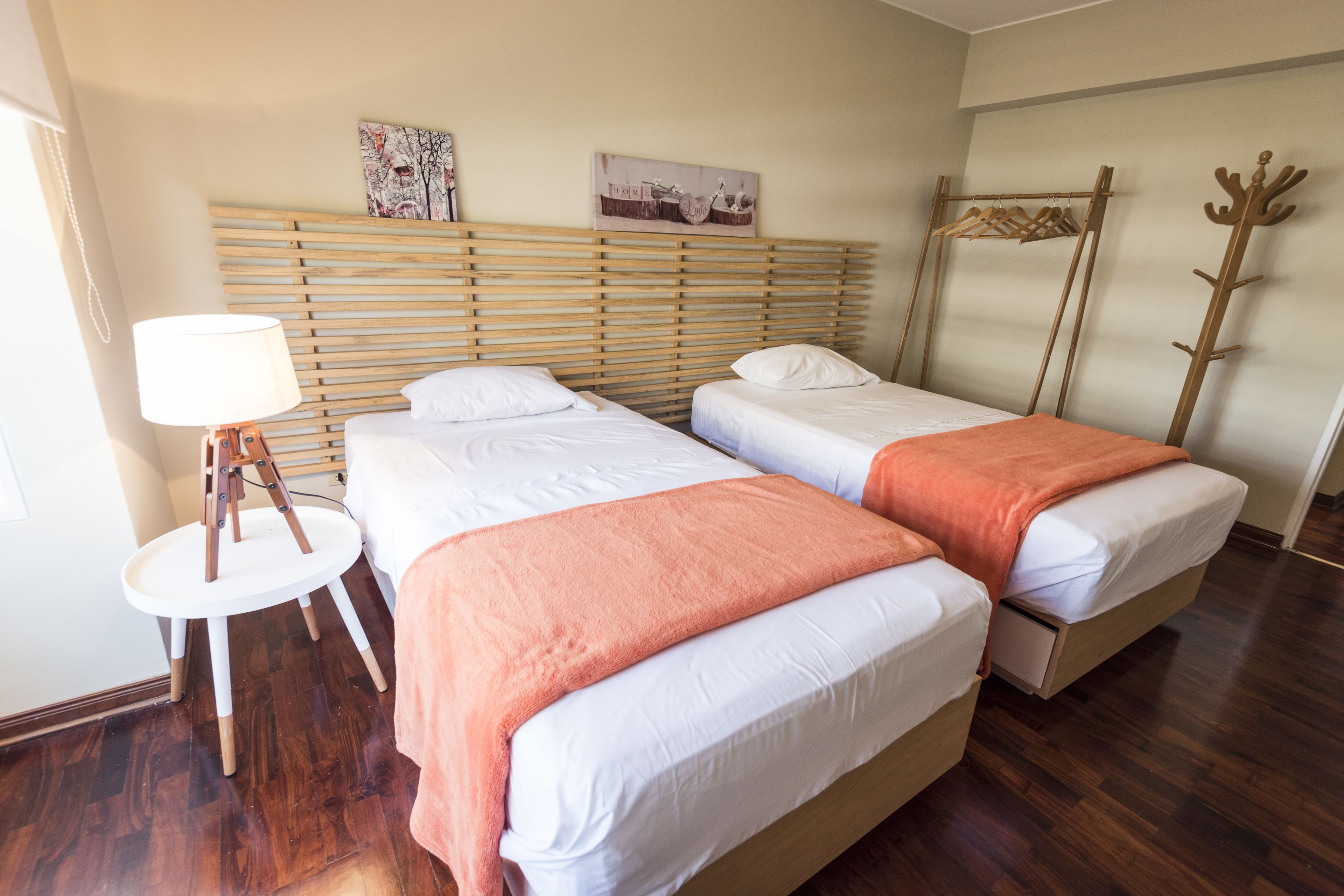 A hotel room with two beds, a wooden headboard, a side table with a lamp, and a coat rack.