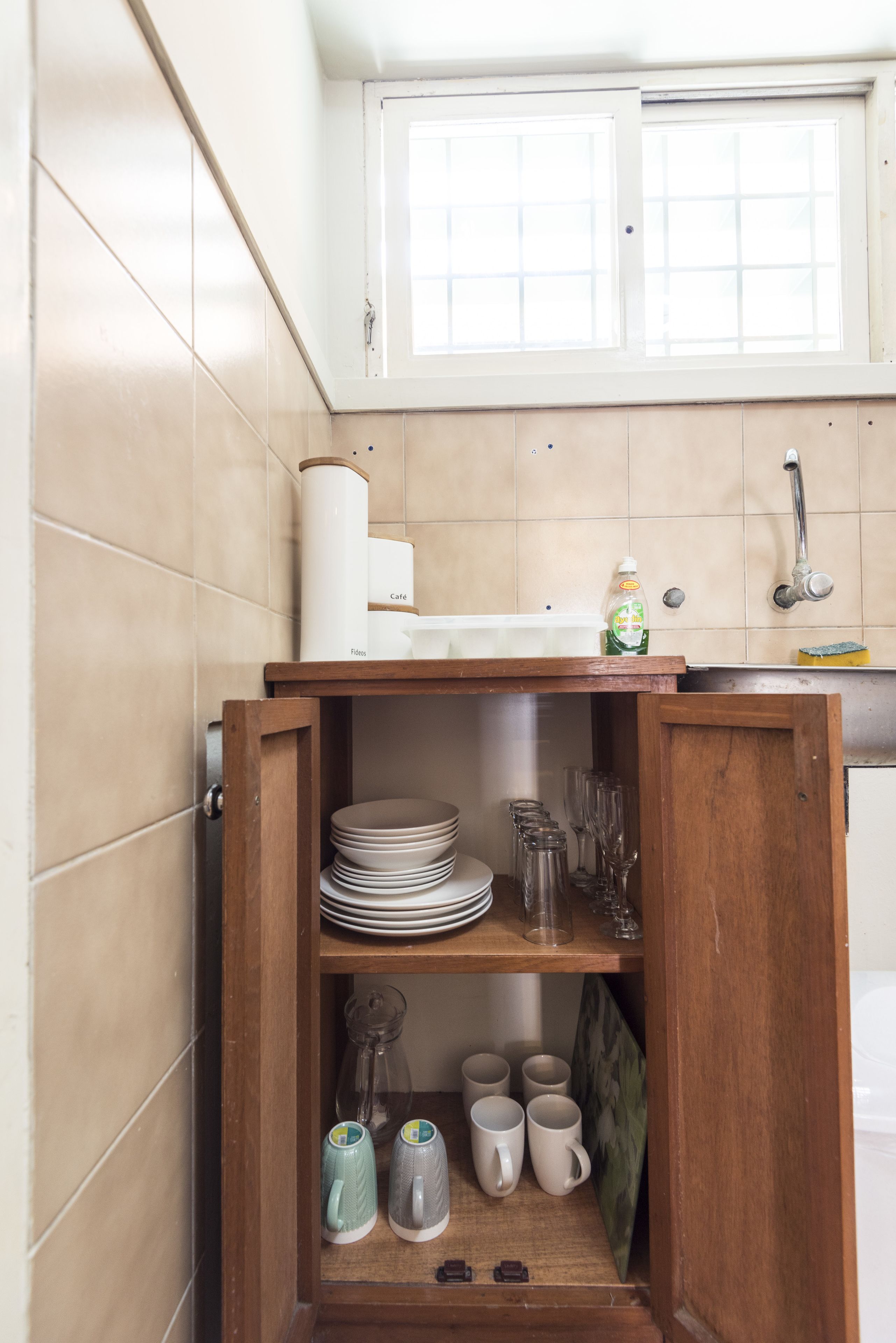 A compact kitchen with a wooden cabinet containing plates, glasses, and mugs. A window above the sink allows natural light to enter.