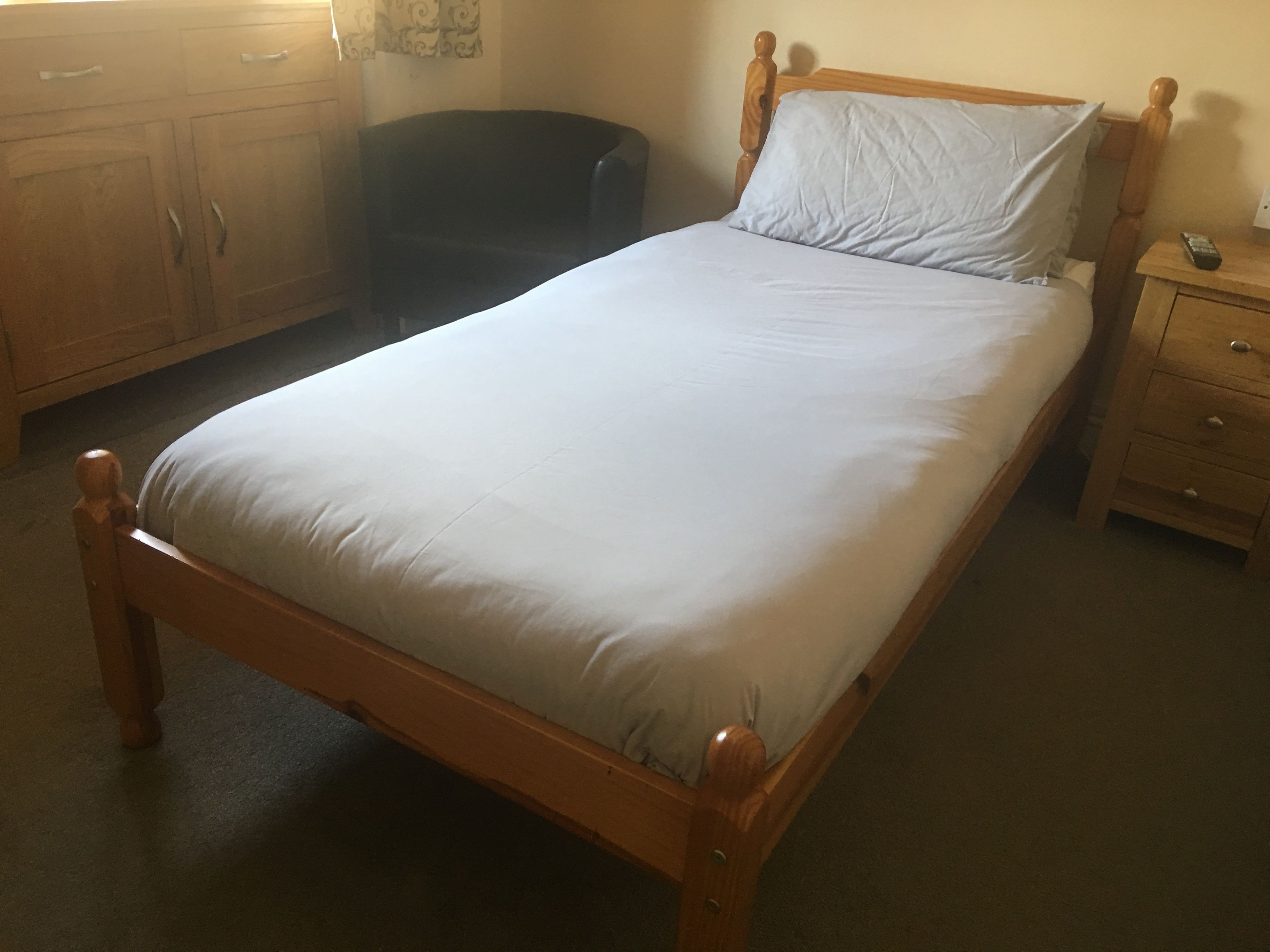 Un letto singolo con struttura in legno e materasso bianco in una stanza con una cassettiera e un comodino in legno.