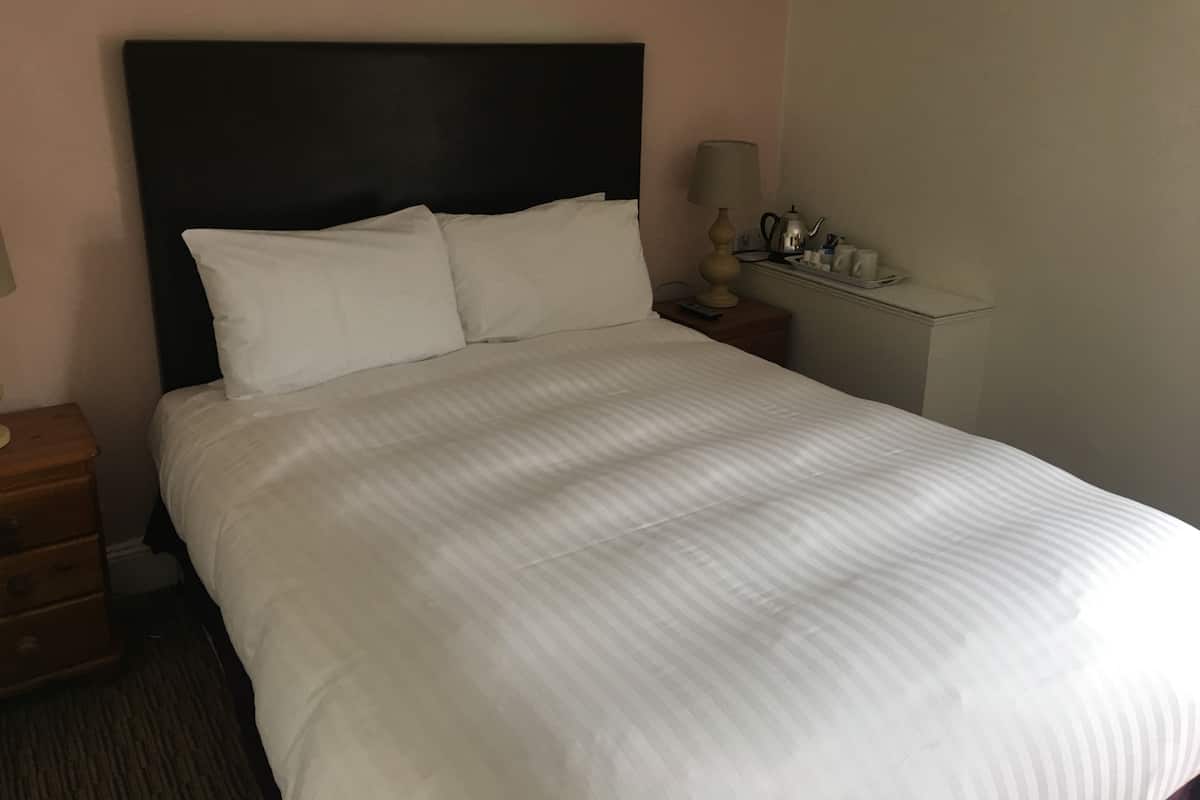 Una cama bien hecha con sábanas blancas, un cabecero oscuro y una mesita de noche con una lámpara y un pequeño neceser.